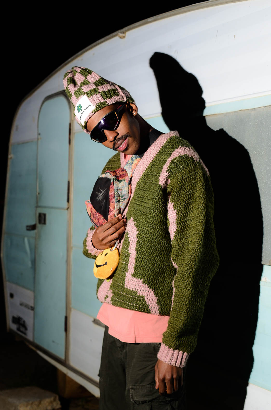 DOOM IT in a green and pink crochet sweater and beanie, leaning against a vintage caravan at night.